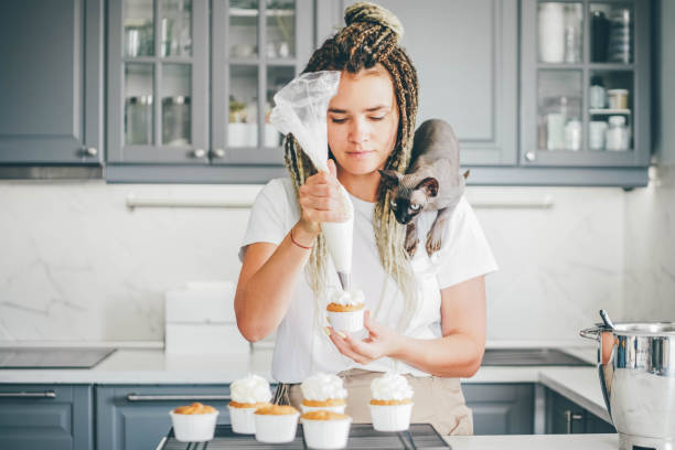 Woman decorating homemade cupcake with cream and cat is watching this process.