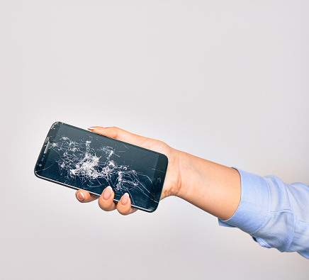 Hand of caucasian young woman holding broken smartphone showing craked screen over isolated white background