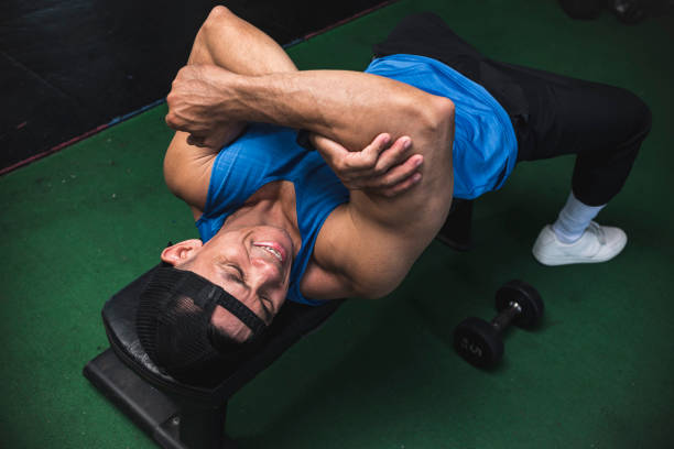 un chico asiático en forma sufre de un tendón del bíceps desgarrado o hiperextendido en un banco mientras hace ejercicio. lesión común de entrenamiento de brazos. - cramping fotografías e imágenes de stock