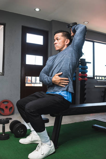 istock A fit athletic asian guy in a sweatshirt does seated overhead dumbbell one arm tricep extensions. Working out and training triceps. Open air gym setting. 1338889110