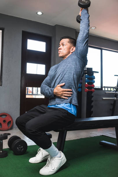 istock A fit athletic asian guy in a sweatshirt does seated overhead dumbbell one arm tricep extensions. Working out and training triceps. Open air gym setting. 1338889124