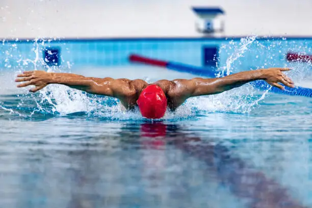 Photo of Professional male swimmer in swimming cap and goggles in motion and action during training at pool, indoors. Healthy lifestyle, power, energy, sports movement concept