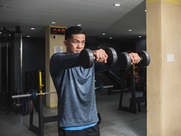 istock A fit asian man in a sweatshirt does double front dumbbell raises. Shoulder workout and training. Open air gym setting. 1338889951