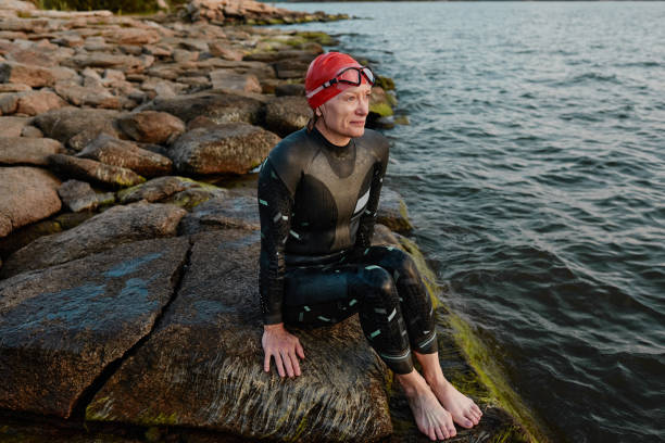 Swimmer sitting on the lakeshore Mature woman in swimsuit sitting on the rock and resting after swimming near the lakeshore wetsuit stock pictures, royalty-free photos & images