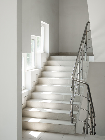 Sun shines through windows on deserted steps of white staircase. Public place inside apartment building. Interior design of urban architecture.