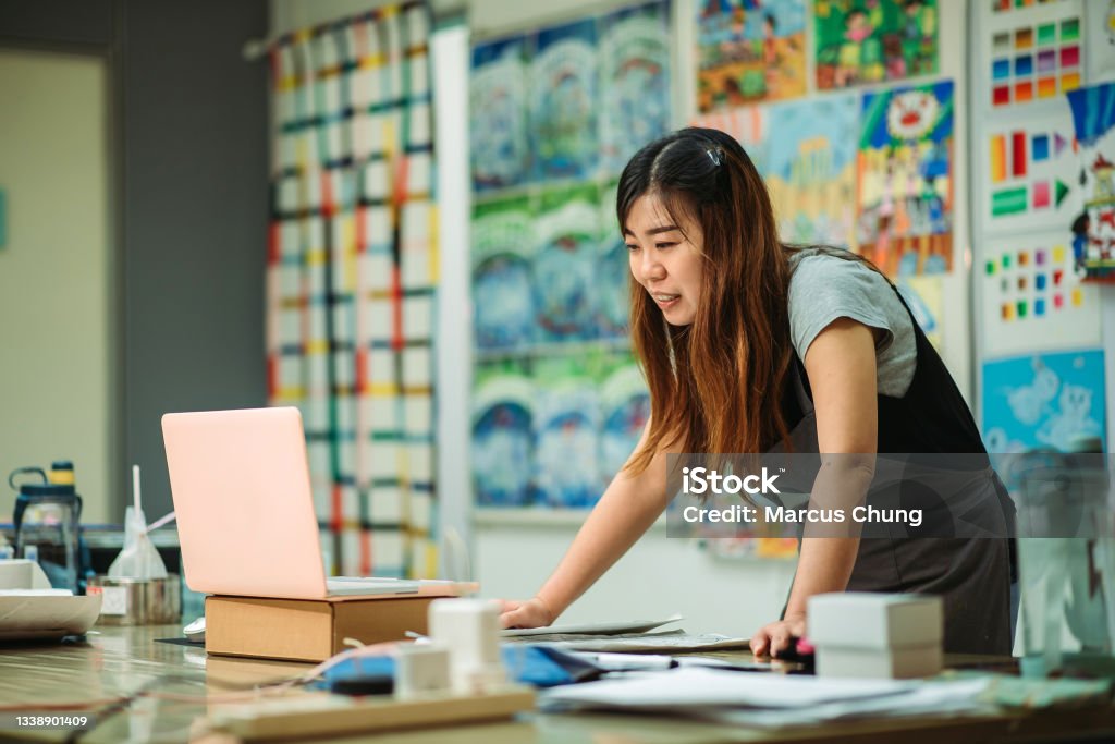 Asian chinese female smiling teacher teaching online art class at art school wide shot of women explaining and talking with students thru laptop during online art class Teacher Stock Photo
