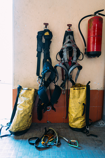 Used firefighter safety outfit with belts and big yellow dirty bags near wall with red fire extinguisher in room with blue floor. Firefighter work tools concept