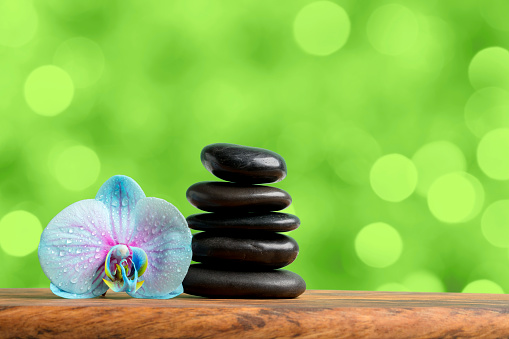 Orchid flower with a stack of black massage stones on a wooden table (mango wood).  Beautiful green bokeh background. Orchid flower is covered with water drops. Space for copy.