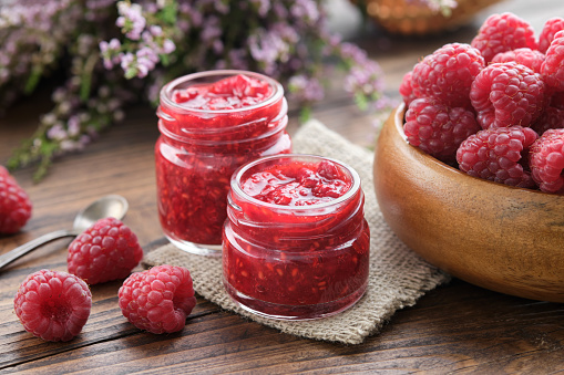 Two small jars of raspberries jam, wooden bowl of fresh red raspberries on kitchen table.