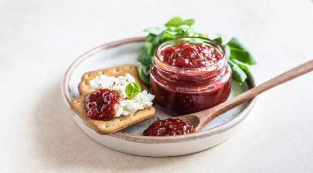 Tomato jam, confiture or sauce in glass jar with crackers and green leaves salad. Unusual savory jam. Mediterranean cuisine. Selective focus.