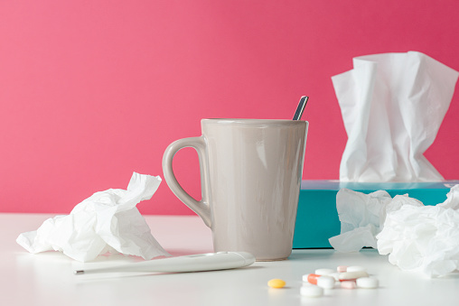 Tissue box, tissues,  thermometer, protective face mask, capsules and a hot drink on white table in front of pink wall.