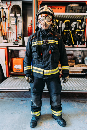 Serious firefighter in protective clothes and heat resistant gloves with helmet on head standing on cement floor behind fire truck with professional instruments. Risk professions concept