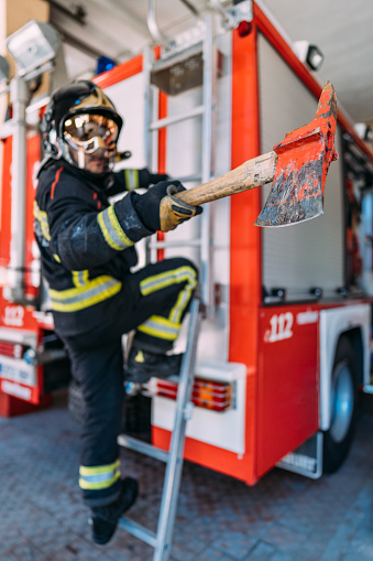 Side view of unrecognizable firefighter in uniform and hardhat carrying big used pickax with wooden handle while standing near fire truck with professional tools and safety vests. Fire extinguishing concept