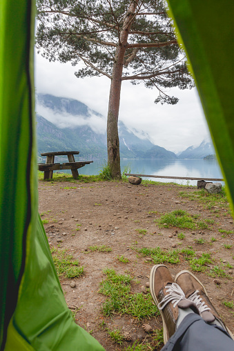Man relaxes in tent after hike and looks on lake and camp bench between pine trees. Exploring north nature at summertime. Lysefjord, Norway.