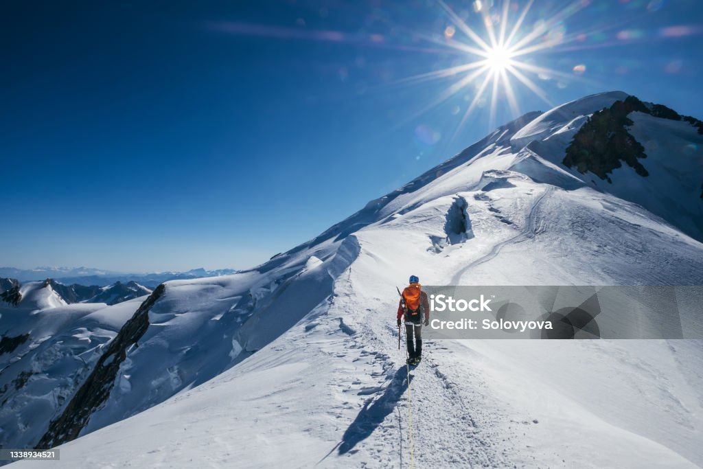 Before Mont Blanc (Monte Bianco) summit 4808m last ascending. Team roping up Man with climbing axe dressed high altitude mountaineering clothes with backpack walking by snowy slopes with blue sky. Mountain Stock Photo