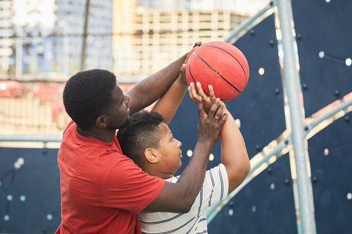 African American father in orange tshirt adjusting hand position of son while teaching him to throw basketball in hoop