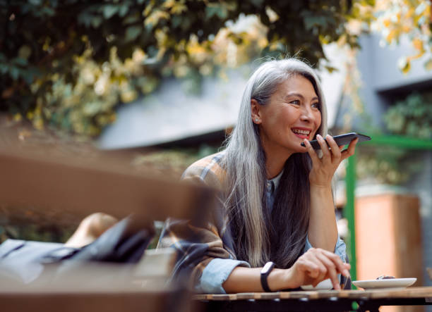 istock Smiling silver haired Asian lady uses loudspeaker mode on smartphone on outdoors cafe terrace 1338936823