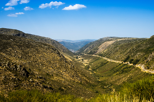 View along the glaciated valley running from the Serra da Estrella mountain range to the town of Manteigas in Portugal. This is a remarkably typical U-shaped glaciated valley that one might expect to find in a geography text book. A small road runs along the valley on the right hand side of the picture and the town of Manteigas is partially visible at the end of the valley. The Zezere river runs along the base of the valley. Deep blue sky with some small clouds overhead.