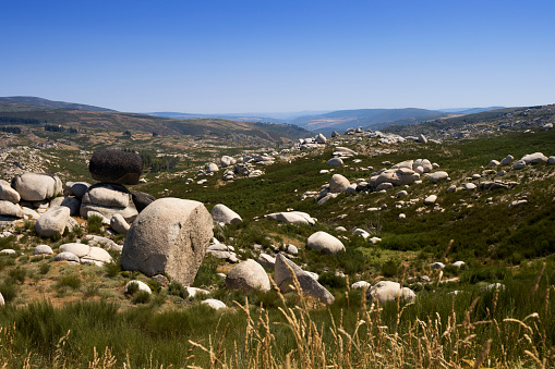 Rock strewn landscape in the Serra da Estrela mountain range of Portugal. Large boulders in the foreground with mountains in the background and a valley in the middle distance. Clear blue sky overhead.