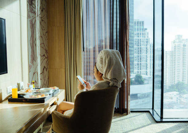 Woman sitting holding coffee cup and using phone Asian woman in a bathrobe and a headscarf is preparing for breakfast after using the phone in her hotel room. room service stock pictures, royalty-free photos & images