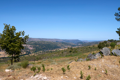 View of the landscape in the Serra da Estrela mountain range of Portugal. A tree left foreground, boulders to the right, mountains in the background and a valley in the middle distance. Clear blue sky overhead. Wide angle view.