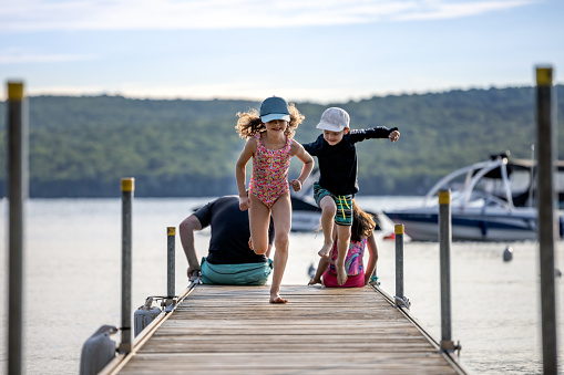 Father with Three Kids Sitting on Pier at the Lake During Summer Vacations, Lac St-Joseph, Quebec, Canada. The single parent is with his son and two daughters. They are looking at the view. Two of the kids are running on the pier and having fun.