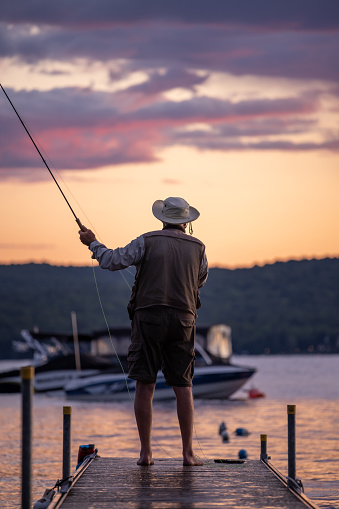 Senior Man Fly-Fishing at Sunset at the lake. He is standing on a pier at Lac St-Joseph, Quebec, Canada.