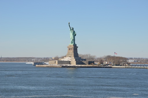 The iconic Statue of Liberty stands tall and proud at the mouth of the Hudson River against the backdrop of a sunny blue sky.