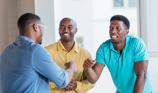 A group of three African-American men standing face to face indoors, smiling and conversing. Two of them are bumping fists. They are mid adult men in their 30s.