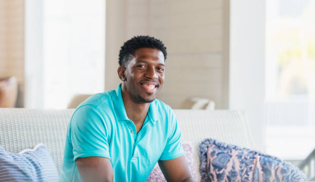 hombre afroamericano sentado en el sofá, sonriendo a la cámara - polo shirt african ethnicity men african descent fotografías e imágenes de stock