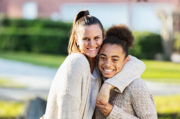 A mixed race 13 year old teenage girl standing in the front yard with her step-mother, arms around each other, holding hands, smiling at the camera.