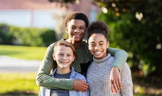 A group of three multi-ethnic children from a blended family standing together outdoors in their yard, posing and smiling for the camera. The caucasian boy on the left is 11 years old. His stepsister and stepbrother are 13 year old twins, mixed race African-American and Caucasian.