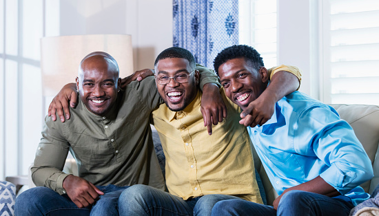 A group of three mid adult African-American men in their 30s hanging out together on a living room couch. They are sitting side by side, arms around one another's shoulders, looking at the camera, smiling and laughing.