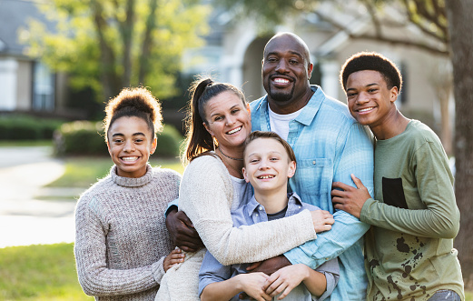 Portrait of a multi-ethnic blended family of five standing together outdoors in their front yard, posing for the camera. The African-American father is in his 40s. Their son and daughter from his prior marriage are standing on the ends. They are 13 year old twins, mixed race African-American and Caucasian. The mother is in her 30s and their youngest son from her prior marriage is 11 years old.
