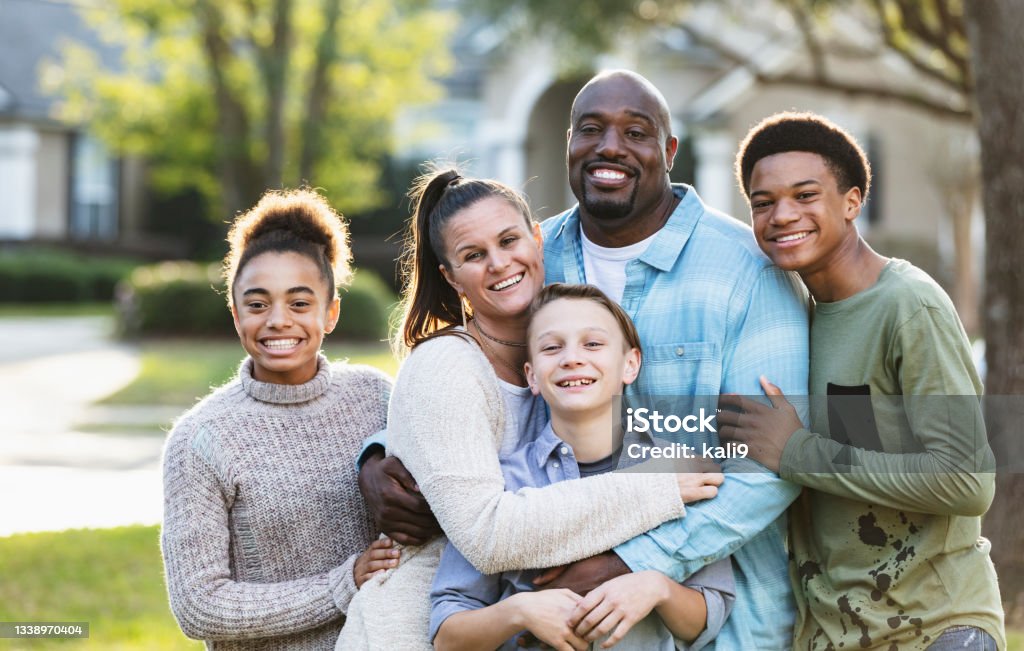 Portrait of blended multi-ethnic family, three children Portrait of a multi-ethnic blended family of five standing together outdoors in their front yard, posing for the camera. The African-American father is in his 40s. Their son and daughter from his prior marriage are standing on the ends. They are 13 year old twins, mixed race African-American and Caucasian. The mother is in her 30s and their youngest son from her prior marriage is 11 years old. Adoption Stock Photo