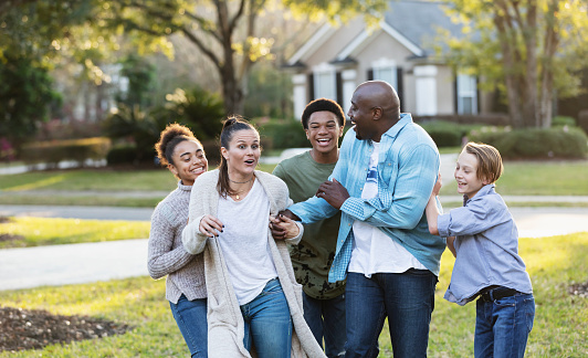 A multi-ethnic blended family of five, with three children, standing together in their front yard, laughing. The African-American father, in his 40s, is holding his wife's hand. She is in her 30s. Their son and daughter from his prior marriage are 13 year old twins, mixed race African-American and Caucasian. Their youngest son from her prior marriage is 11 years old.
