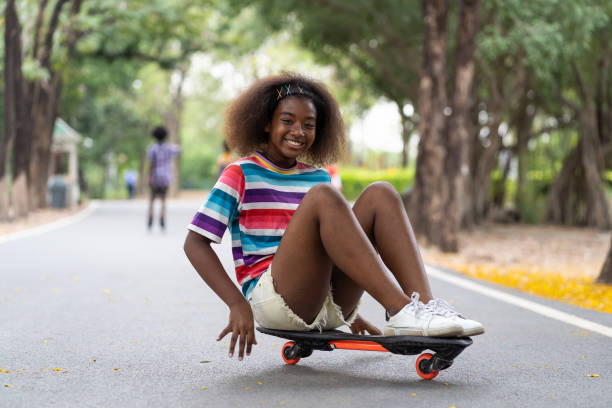 uma jovem feliz brincando no skate no parque. garota afro-americana com cabelo encaracolado praticando skate no jardim. - skateboard park fotos - fotografias e filmes do acervo