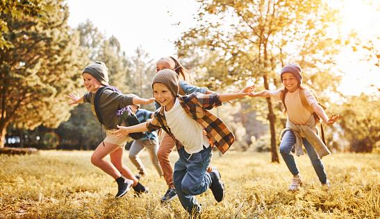 Group of happy joyful school kids with backpacks running with outstretched arms in forest on sunny spring day, excited children scouts boys and girls having fun during camping activity in nature