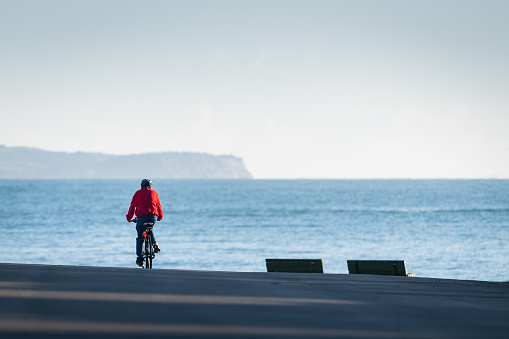 A cyclist riding towards beach with a backdrop of distant hills, Milford beach, Auckland.