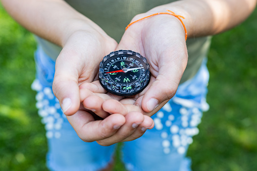 Girl holding the compass. Hands of a teenager holding a liquid compass. Red compass needle points north. Green grass background. Copy space. Orienteering on the ground. Selecting the direction