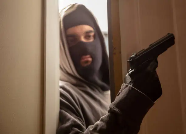 Controlling the area before the invasion. Blur armed young man in grey hoodie and full face masked behind half opened door. Robber criminal with black balaclava leather glove and pistol, gun, handgun.