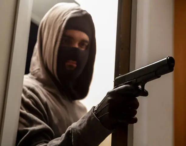 Ready for invasion concept. Thief robber killer burglar gangster terrorist armed young man in grey hoodie with black balaclava holding pistol in leather glove behind half opened door. Target attack.