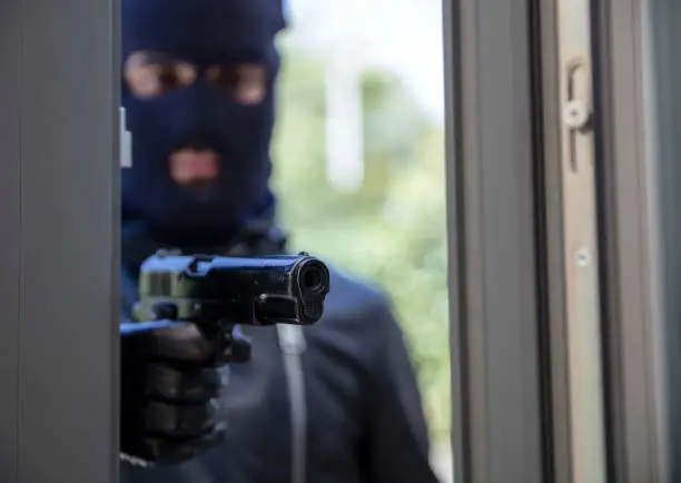 Killer with balaclava concept. Man in black hood holds gun in leather gloved hand ready to spread fear and violence. Blur armed robber aims with pistol behind window. Action plan, terrorism, burglary