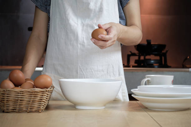 istock Female cook in a white apron is cracking an egg in home's kitchen. 1338996595