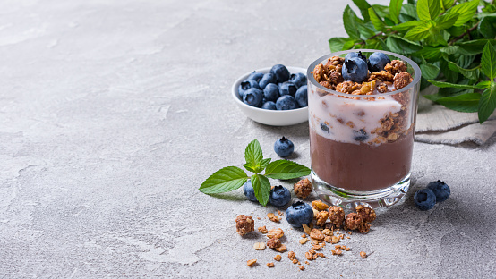 Tasty chocolate pudding or sweet mousse with yogurt, blueberry, granola and mint in portion glass on grey concrete background