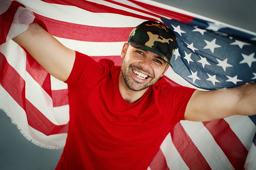 Closeup front view of a cheerful 30's guy holding American flag and looking at the camera.
