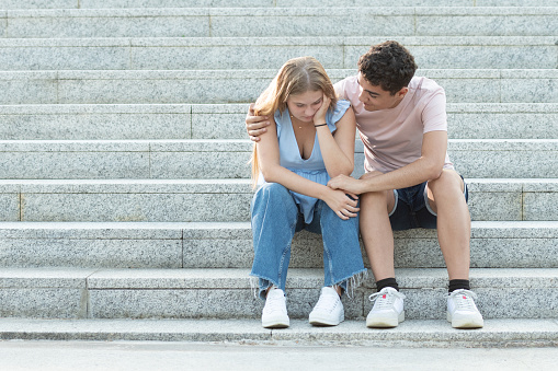 Teenager couple sitting on the stairs. Hispanic boyfriend trying to cheer up and hugging sad girlfriend. Relationship troubles, anxiety and depression in adolescence concept with copy space.