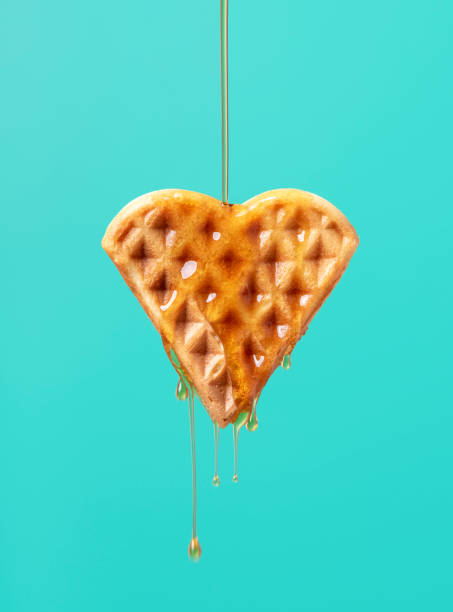 Waffle with maple syrup isolated on a green background. Pouring maple syrup over one heart-shaped waffle against a green-colored background. Close-up of maple syrup dripping on a waffle. waffle stock pictures, royalty-free photos & images