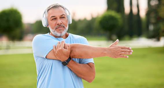 Positive active senior sportsman wearing wireless headphones doing arm stretching, warming up muscles before jogging or outdoor workout, mature man exercising outside in city park in morning
