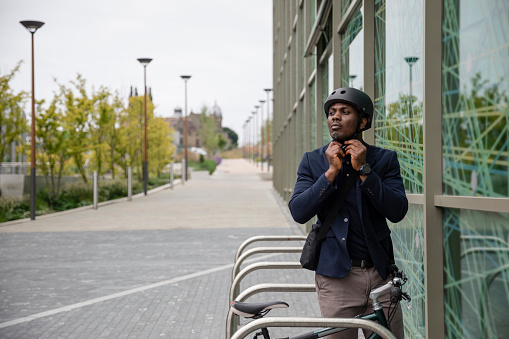 A mature black man wearing formal businesswear and a helmet on a summers day in a city. He is arriving at work with his bicycle and removing his helmet after sustainably commuting to work.